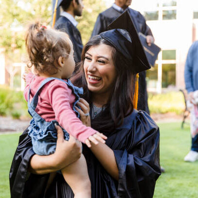 Young mother, graduation gown, smiling, holds toddler. Outdoors, family celebration.