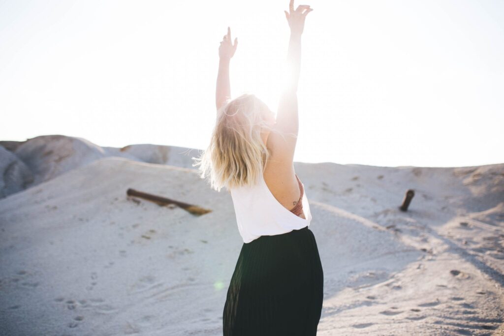 woman on a dune with both arms raised toward the sun and sky