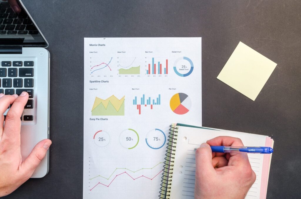 hands writing in a notebook and typing on a laptop over charts and graphs on a desk